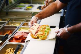 A person assembling a sub sandwich at a deli counter surrounded by ingredients in containers