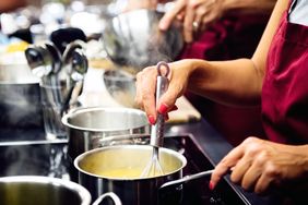 A person using a whisk to stir contents in a pot on a stove wearing an apron over their clothing with culinary utensils nearby