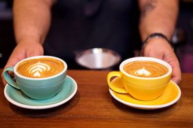Two cups of coffee with latte art designs on a counter one in a green cup and the other in a yellow cup hands placing them down