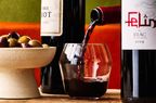 A glass being filled with red wine with bottles and a bowl of olives on a table