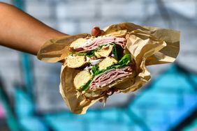 A bagel sandwich cut in half with deli meats and lettuce held in a persons hand wrapped in paper