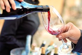 Person pouring red wine into a glass several wine bottles visible in the background