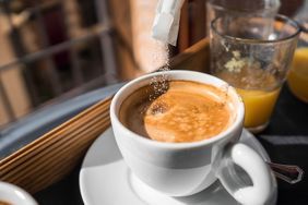 A cup of coffee with sugar being added alongside a glass of juice in the background