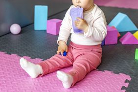 A baby seated on a play mat holding a squeezable pouch with blocks in the background