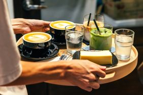 A person carrying a tray with various drinks and a dessert