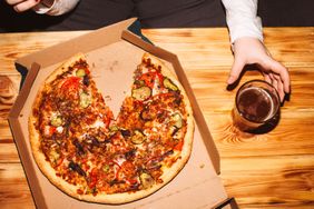 A pizza in an open box on a wooden table a person reaching for a beverage nearby