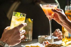Hands holding cocktail glasses in a toast gesture at a table setting with snacks