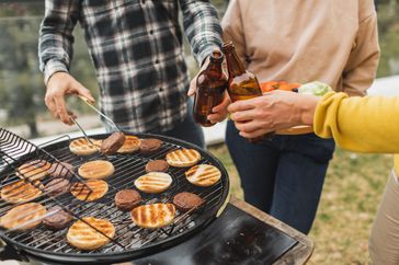 People around a barbecue grill with burgers and buns holding drinks and cooking together outdoors