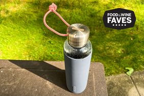 A reusable glass water bottle with a metal cap and a red carrying loop labeled blackblum set on a table outdoors