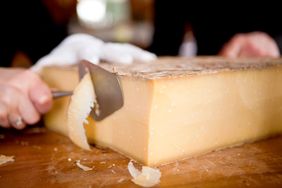 A person slicing cheese with a cheese knife on a wooden counter