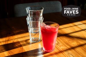 A glass of red juice with ice next to two stacked empty glasses on a wooden table with a Food Wine Faves label in the corner