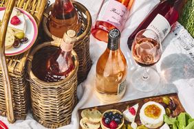 A picnic setup featuring bottles of ros wine a wine glass wicker baskets and a spread of fruits bread and cheese
