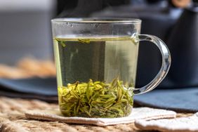 A glass mug with loose green tea leaves.