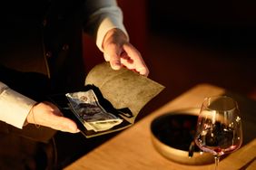 Server holding a bill folder with money over a dining table a glass of wine and a bowl visible