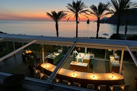 A restaurant with glass walls people inside preparing food overlooking a scenic coastline at sunset with palm trees and the sea