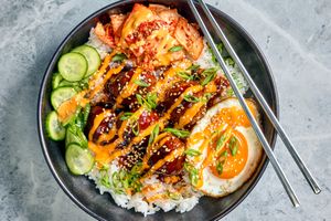 A bowl of rice topped with Korean meatballs a fried egg kimchi cucumbers and garnished with sauce and green onions