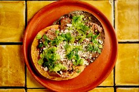 Two loaded tacos on a clay plate with toppings such as meat cheese guacamole and herbs