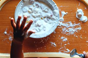 Childs hand reaching for a bowl of yogurt with scattered food on a wooden surface