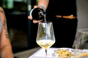 A person pouring white wine into a glass with snacks and a table in the foreground