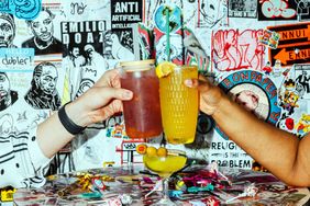 Two hands clinking cocktail glasses in a vibrant restaurant with graffiticovered walls and drinks on the table