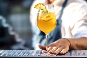 A summer cocktail in a stemmed glass garnished with lemon and lime slices placed on a bar table with a person in the background