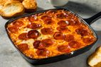 A skillet filled with a baked pepperoni topping on a food dish accompanied by slices of bread