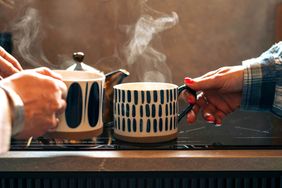 Two hands holding mugs with steam rising a steaming kettle in the background
