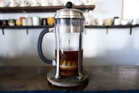 A French press with brewed coffee on a table with shelves of mugs and cups in the background