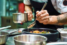 A chef plating food in a kitchen using a spoon and sauce pan