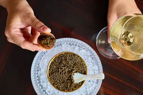 Serving of caviar in a bowl on ice with a glass of champagne and a hand holding a portion on a cracker