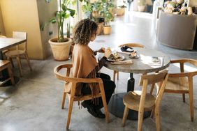 A person eating alone at a small table in a restaurant, with other tables and plants visible in the background