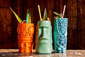 Three tiki-style mugs containing garnished beverages on a textured table