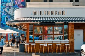 Exterior view of Milkbread cafe with people walking outside featuring stools and a counter under a canopy