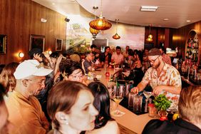 A bartender prepares drinks in a crowded bar with people seated and standing around the counter