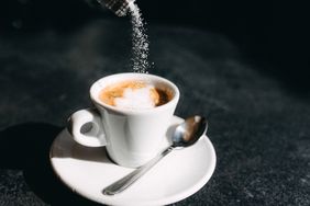 Sugar being poured into a cup of espresso on a saucer with a spoon placed beside it